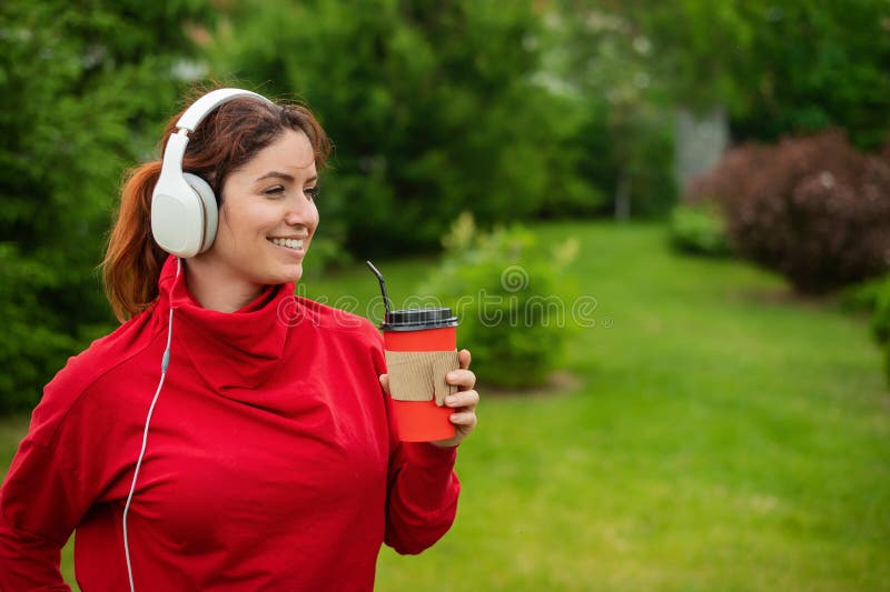 Red-haired woman listens to music on headphones and drinks coffee with a straw in the park. A girl enjoys a walk on a