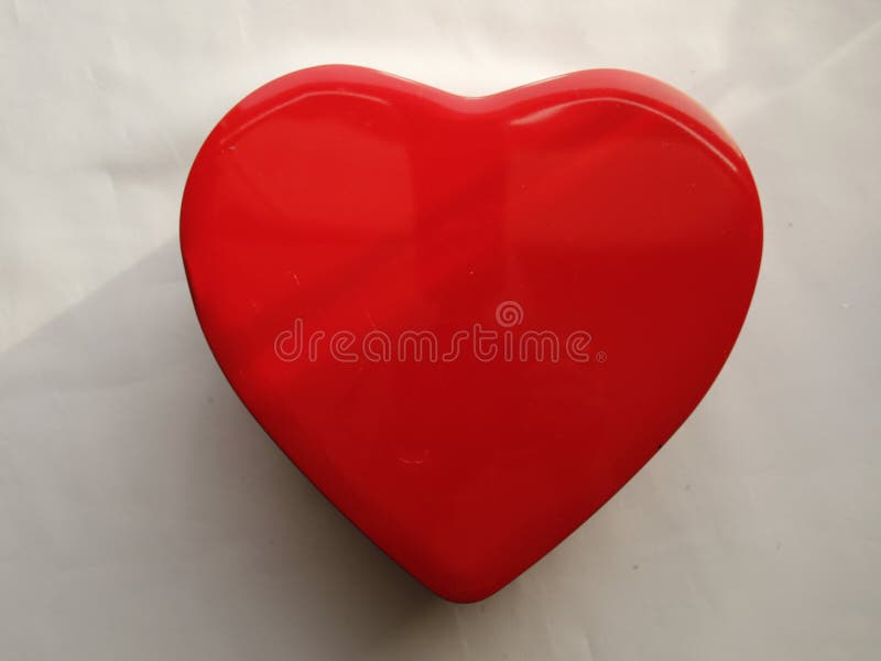 Red shiny heart shape metal tin - LOVE. Red heart-shaped metal tin with a glossy finish, resting on a white surface. Its smooth, shiny surface reflects light, enhancing its vibrant color and curved design. The tin appears compact, suitable for storing small items or as a decorative piece.