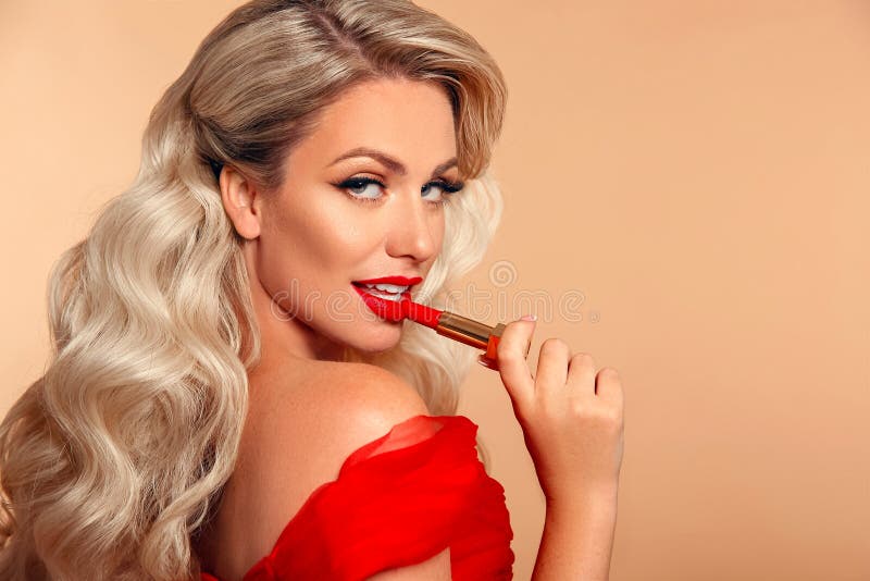 Red lipstick shop. Beauty woman makeup. Fashion glamour portrait of pretty blonde with long wavy hair style applying lipstick over beige background. Beautiful female face. Make up product cosmetics