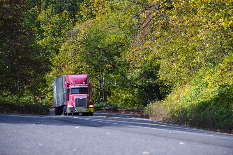 Red modern semi truck with trailer going up hill in autumn trees