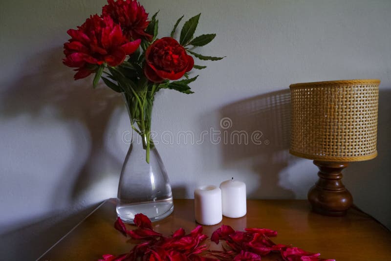 Red peonies in glass vase, straw lamp, white candles on wooden commode across white wall. Interior design. Home decor. lights and shadows concept.