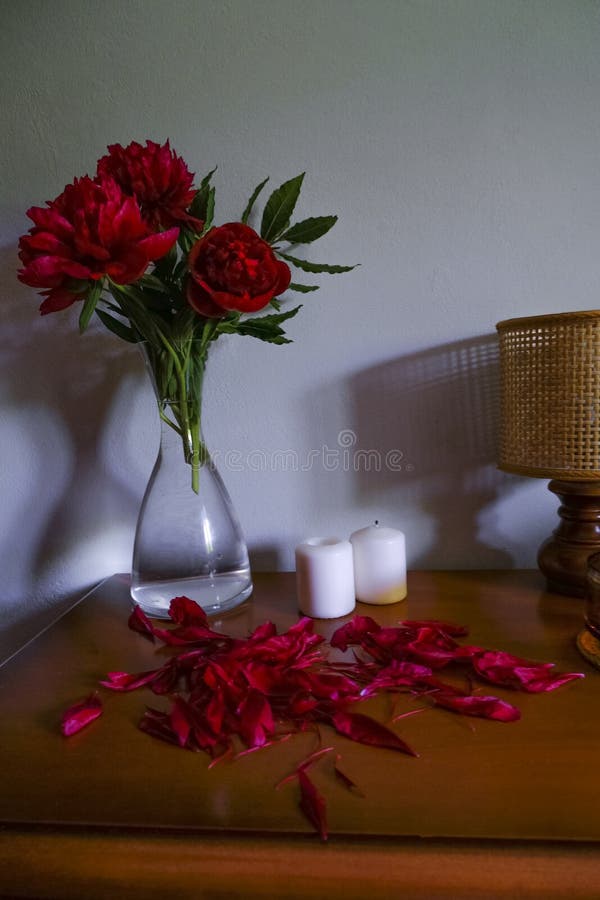 Red peonies in glass vase, straw lamp, white candles on wooden commode across white wall. Interior design. Home decor. lights and shadows concept