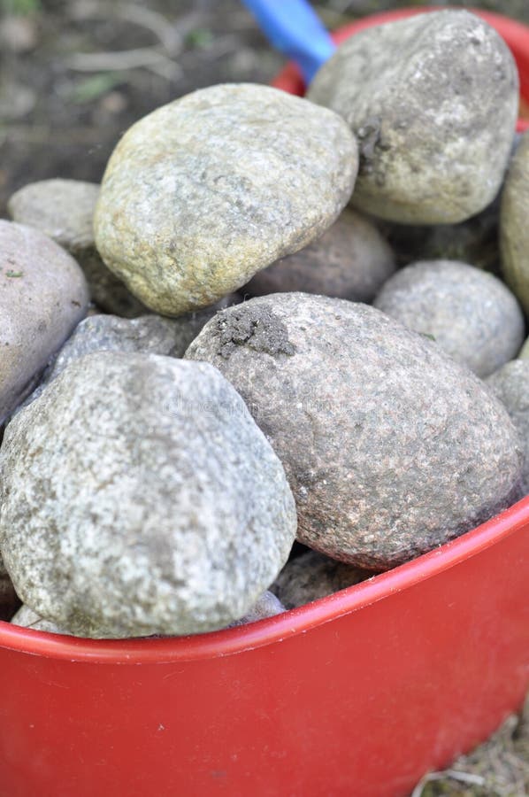 Red plastic bucket full of stones. Round solid granite stones are hard material. Bucketful of muddy rocks