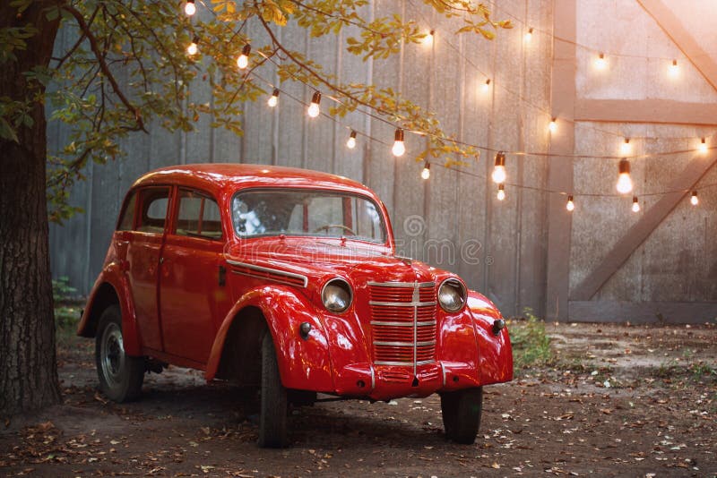 Red retro car standing in the garden in the summer on a background of gray wall and burning bulbs. Vintage vehicle near the garage. Decorated with lights stock image Red retro car standing in the garden in the summer on a background of gray wall and burning bulbs. Vintage vehicle near the garage. Decorated with lights stock image