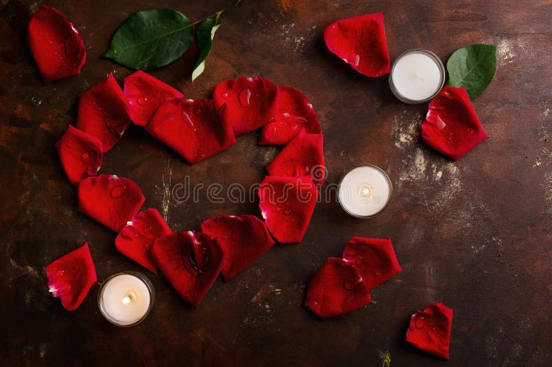 Red rose petals in heart shape with white candles on dark brown and golden background. Love, romance, anniversary, valentine`s day