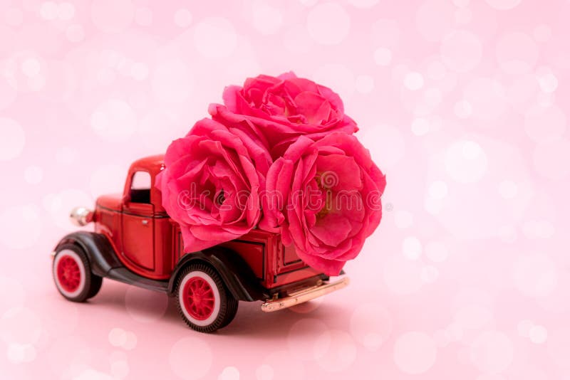 Red toy truck delivering bouquet of rose flowers on pink background with bokeh. February 14, Valentine`s day, 8 March