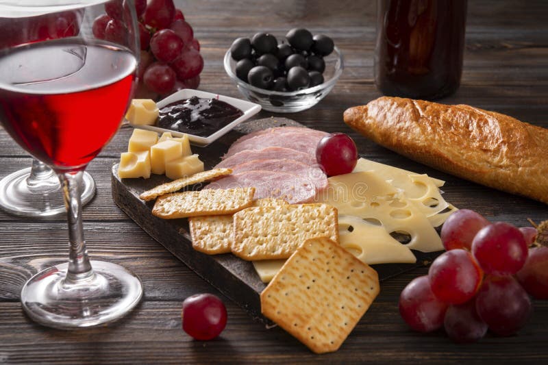 Red wine glasses,  cheese , cracker, ham, olives, jam, grapes, baguette, bottle, corkscrew,  on a dark wooden background