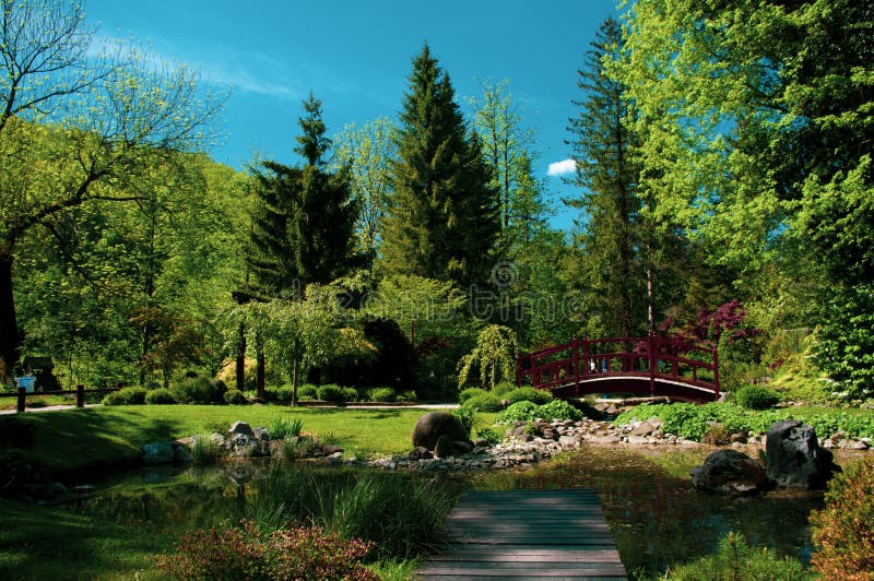 Red wooden bridge over little pond in city park Mozirje, Slovenia. Summer time, green trees and blue sky.