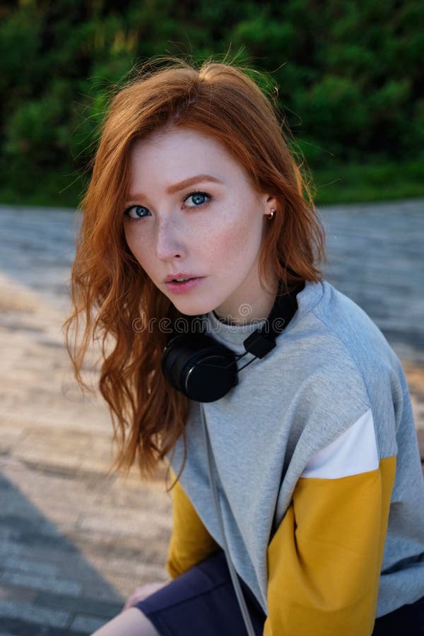 Redhead Girl With Freckles And Blue Eyes Sits In A Park At Sunset And