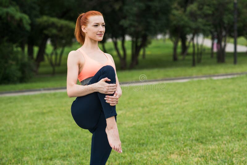 A Redhead Girl Standing On A Mat In The Park And Practicing Yoga Stretching The Leg Stock