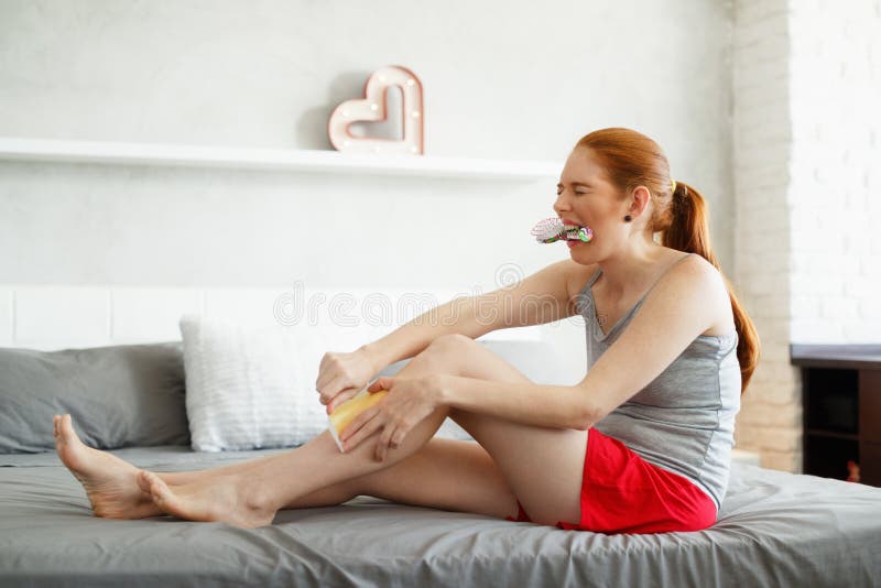 Young woman sitting on bed, doing routine leg waxing. The redhead girl bites a hairbrush to stand the pain while she pulls the strip, then faints back. Young woman sitting on bed, doing routine leg waxing. The redhead girl bites a hairbrush to stand the pain while she pulls the strip, then faints back