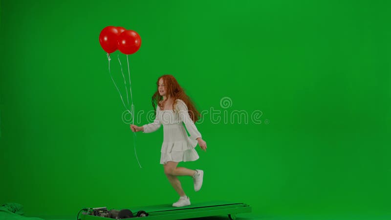 Redheaded little girl in white dress running with red balloons on green background of studio. Half turn. Concept of