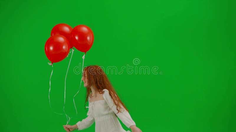 Redheaded little girl in white dress running with red balloons on green background of studio. Half turn. Concept of