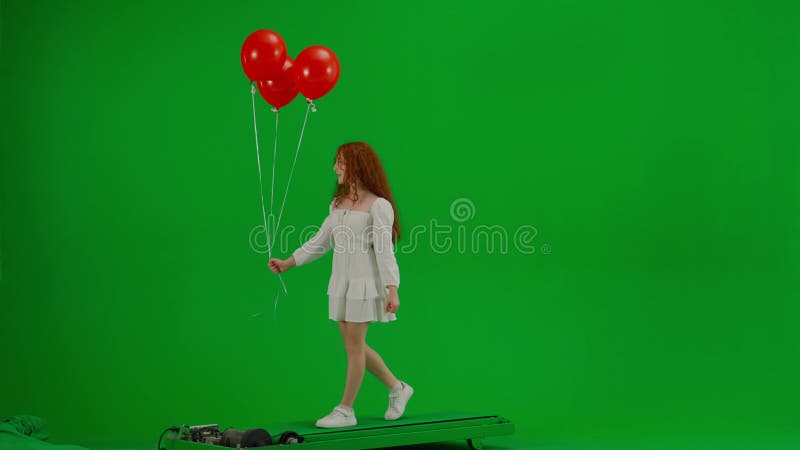 Redheaded little girl in white dress walking with red balloons on green studio background. Half turn. Concept of holiday