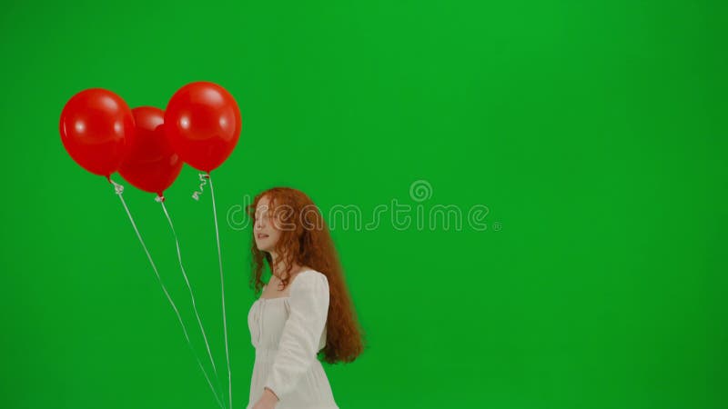 Redheaded little girl in white dress walking with red balloons on green studio background. Half turn. Concept of holiday