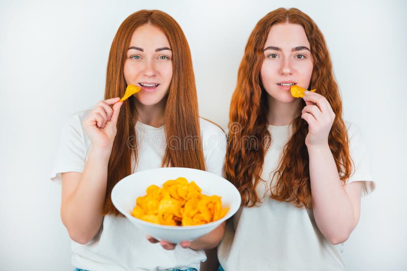 2 Redheaded молодой женщины есть хрустящие картофельные чипсы выглядят счастливыми на изолированной белой концепции высококалорийн стоковые фотографии rf