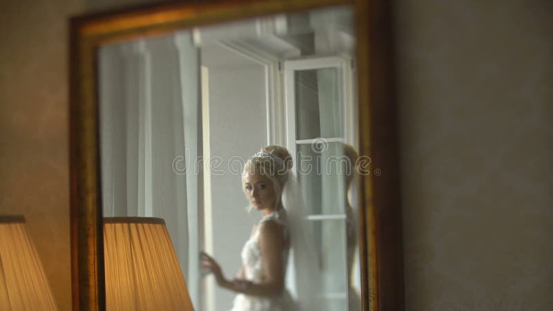 Reflection in the mirror of the adorable blonde bride in the stylish wedding dress, veil and with wonderful make-up