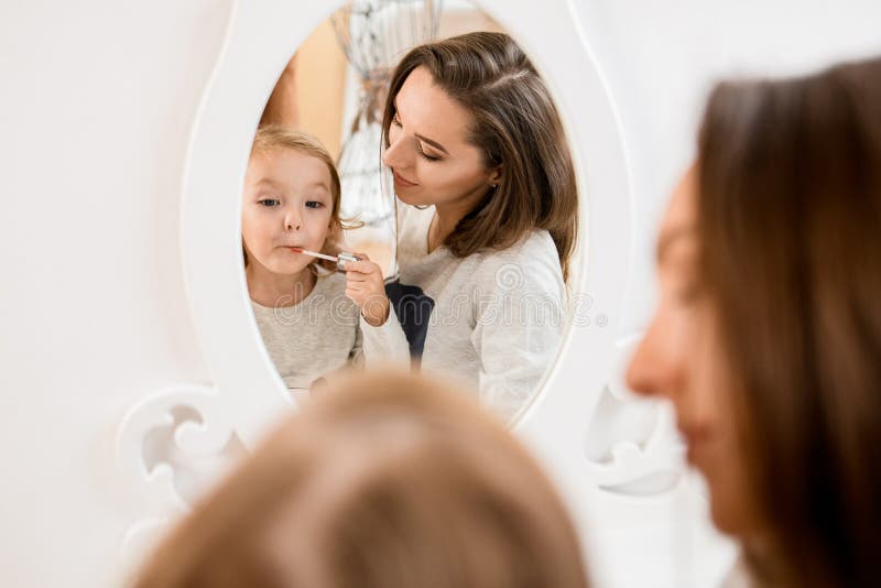 Reflection in mirror of beautiful women and little girl who paints her lips with shiny lip gloss. Reflection in mirror of beautiful women and little girl who paints her lips with shiny lip gloss.