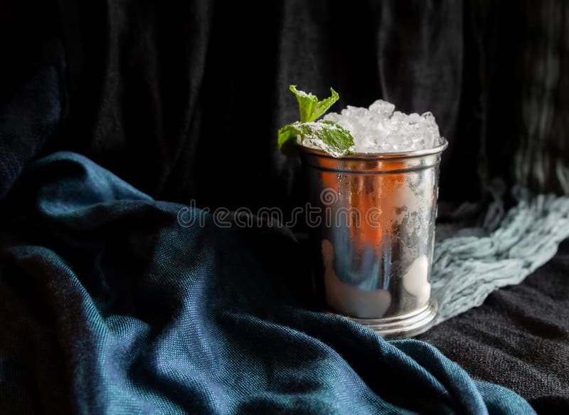 Refreshing mint julep whiskey cocktail or in misted frozen metal glass on dark black fabric drapery background. Bar menu, alcohol recipe decor, specialized website blog design