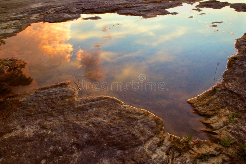 A pool of water trapped in a depression within a rock formation reflects the sky, showing storm clouds rolling in at sunset. A pool of water trapped in a depression within a rock formation reflects the sky, showing storm clouds rolling in at sunset.