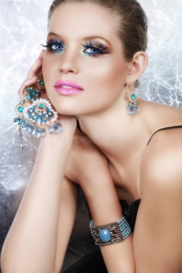 Blond woman with long false lashes holding pearl and blue stones golden jewelry with dreaming expression. Blond woman with long false lashes holding pearl and blue stones golden jewelry with dreaming expression