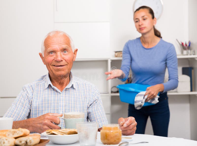 Mature father drinking tea in the kitchen. Adult daughter does cleaning in the apartment. High quality photo. Mature father drinking tea in the kitchen. Adult daughter does cleaning in the apartment. High quality photo