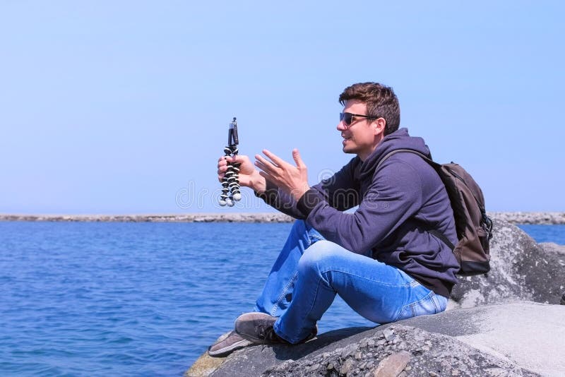 Reise Blogger, den Mann Video auf Smartphone schießt, sitzt auf Stein am Seestrand lizenzfreie stockfotografie