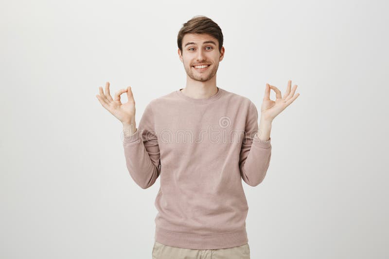 Reizend junger Mann mit hellem Lächeln und Borste, tragender zufälliger Pullover und o.k. darstellen oder Zengeste bei der Stellu lizenzfreie stockfotos Reizend junger Mann mit hellem Lächeln und Borste, tragender zufälliger Pullover und o.k. darstellen oder Zengeste bei der Stellu lizenzfreie stockfotos