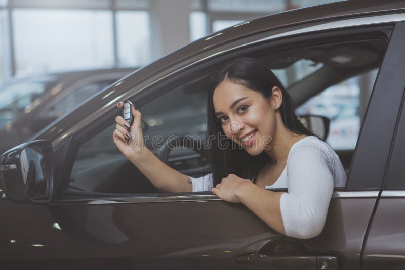 Cheerful beautiful woman smiling happily, holding car key sitting in her new automobile at dealership salon. Lovely female driver buying new car. Ownership, insurance, travelling concept. Cheerful beautiful woman smiling happily, holding car key sitting in her new automobile at dealership salon. Lovely female driver buying new car. Ownership, insurance, travelling concept