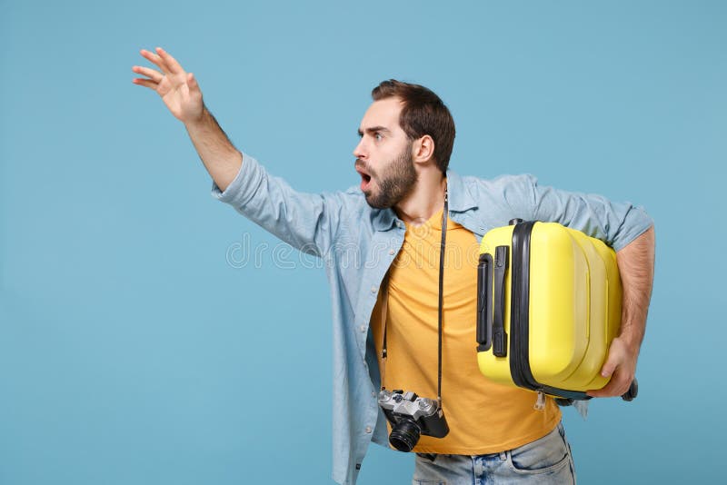 Traveler tourist man in yellow clothes with photo camera isolated on blue background. Male passenger travele abroad on weekends. Air flight journey. Hold suitcase waving hand for greeting, catch taxi. Traveler tourist man in yellow clothes with photo camera isolated on blue background. Male passenger travele abroad on weekends. Air flight journey. Hold suitcase waving hand for greeting, catch taxi