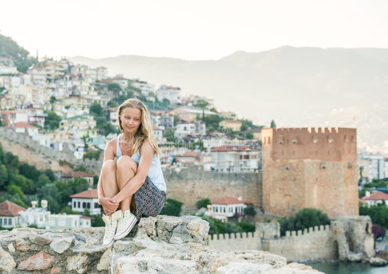 Relajación turística de la mujer rubia joven en la pared antigua de la fortaleza del castillo de Alanya Kizil Kule o torre roja e fotografía de archivo Relajación turística de la mujer rubia joven en la pared antigua de la fortaleza del castillo de Alanya Kizil Kule o torre roja e fotografía de archivo