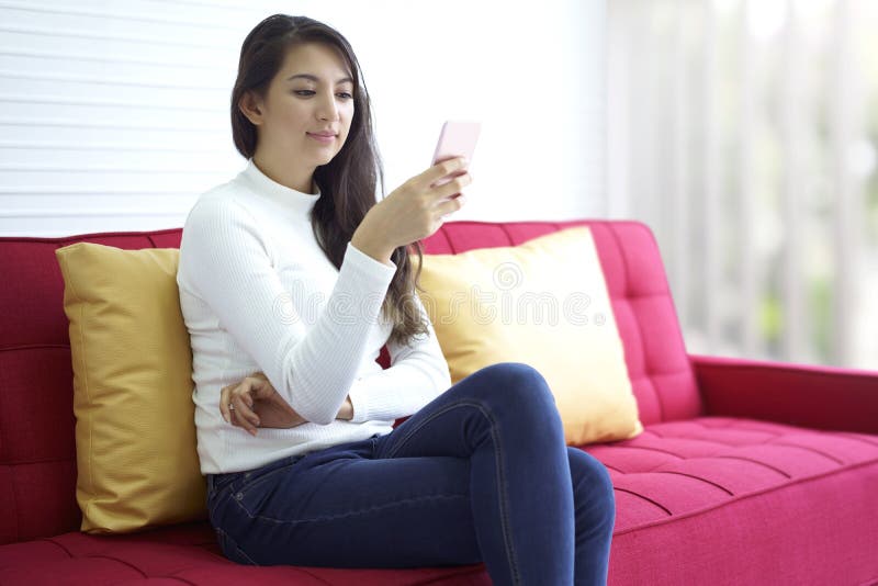 Relaksująca piękna dziewczyna szczęśliwa i używa mądrze telefon podczas gdy siedzący spoczynkowy wygodny na czerwonej kanapie w d obrazy stock Relaksująca piękna dziewczyna szczęśliwa i używa mądrze telefon podczas gdy siedzący spoczynkowy wygodny na czerwonej kanapie w d obrazy stock