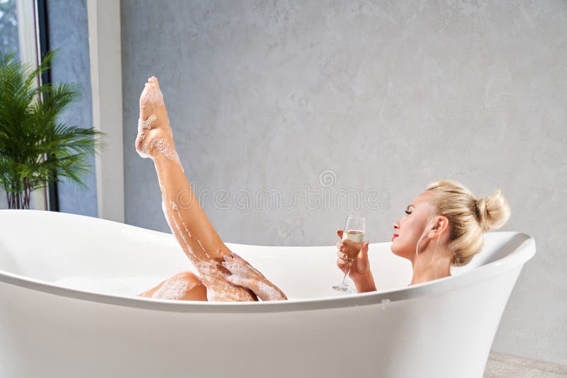 Relaxed blonde woman with hair bun, taking warm bath at home in evening. Side view of female lying in bathtub, with leg sticking out of bubble bath, in front of grey wall. Concept of spa and relax. Relaxed blonde woman with hair bun, taking warm bath at home in evening. Side view of female lying in bathtub, with leg sticking out of bubble bath, in front of grey wall. Concept of spa and relax.