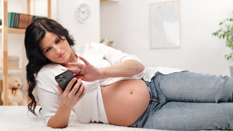 Relaxed casual brunette zwangere vrouw die op internet surft gebruik smartphone applicatie die op bed ligt