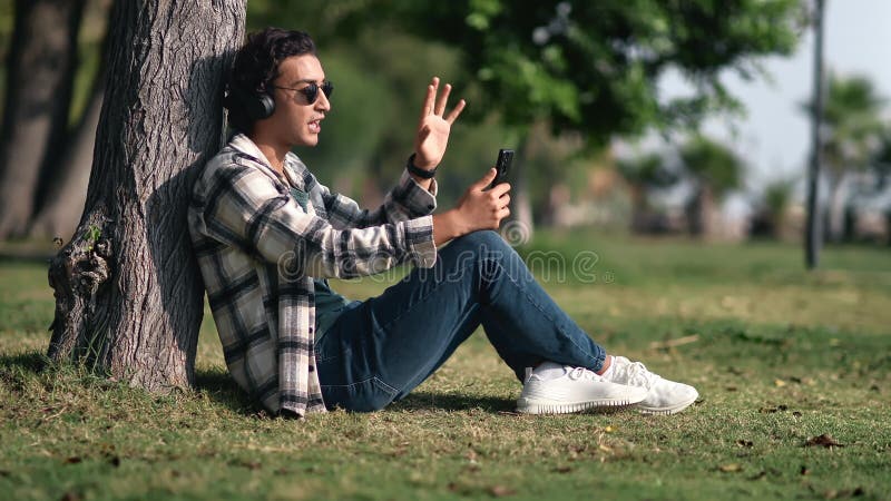 Relaxed Hispanic modern guy talking online video call use smartphone headphones at summer park stock video