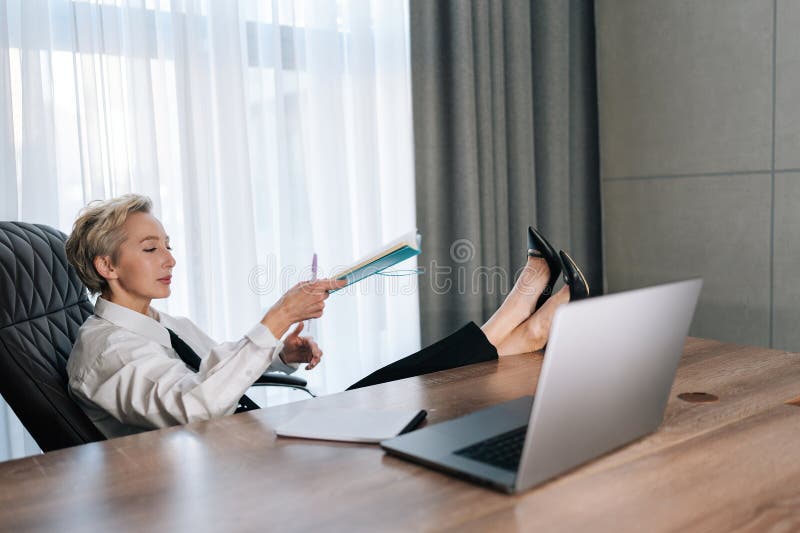 Relaxed middle-aged female CEO putting feet on desk with laptop. Confident successful business woman thinking about career growth relaxing in cabinet. Concept relaxation at workplace