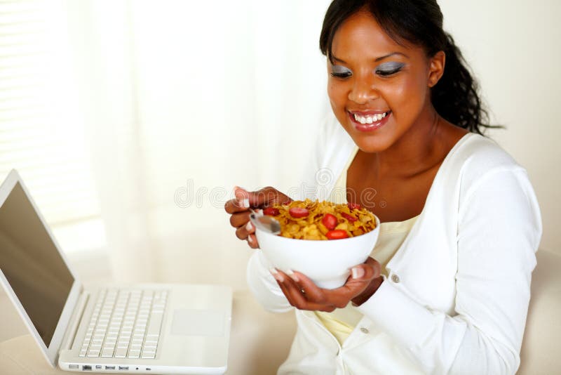 Portrait of a relaxed young woman having healthy breakfast while is sitting on sofa at home in front of her laptop. Portrait of a relaxed young woman having healthy breakfast while is sitting on sofa at home in front of her laptop