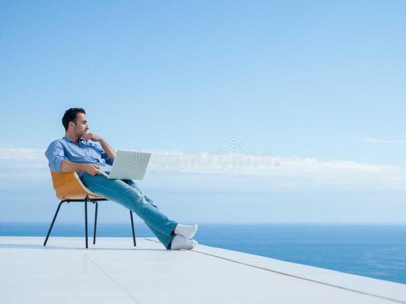 Pohledný mladý muž relaxační a pracuje na přenosný počítač doma balkoně při pohledu na západ slunce.