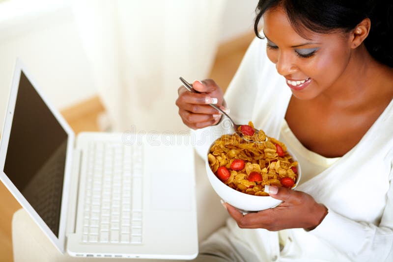 Portrait of a relaxed young woman having healthy breakfast while is sitting on sofa at home in front of her laptop