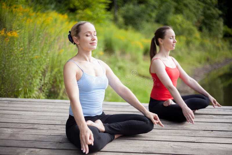 Two serene young beautiful women on meditation session outdoors, sitting cross-legged in Ardha Padmasana Posture, working out in park on summer day, practicing pranayama with closed eyes, full length. Two serene young beautiful women on meditation session outdoors, sitting cross-legged in Ardha Padmasana Posture, working out in park on summer day, practicing pranayama with closed eyes, full length