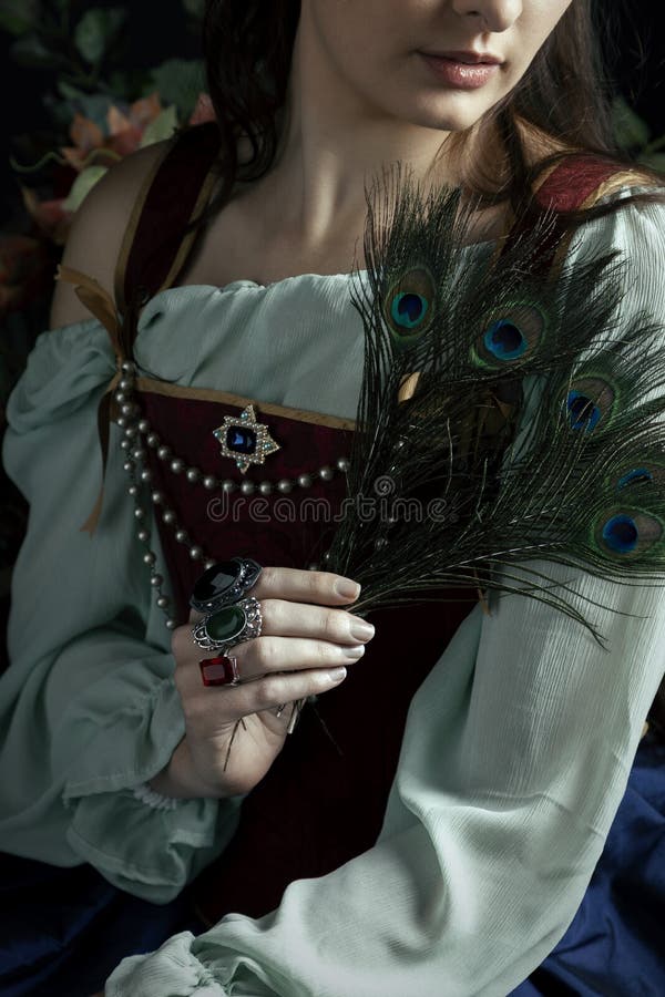 A Renaissance or Fantasy/fairytale/gypsy woman wearing a red brocade corset and blue silk skirt and holding peacock feathers.