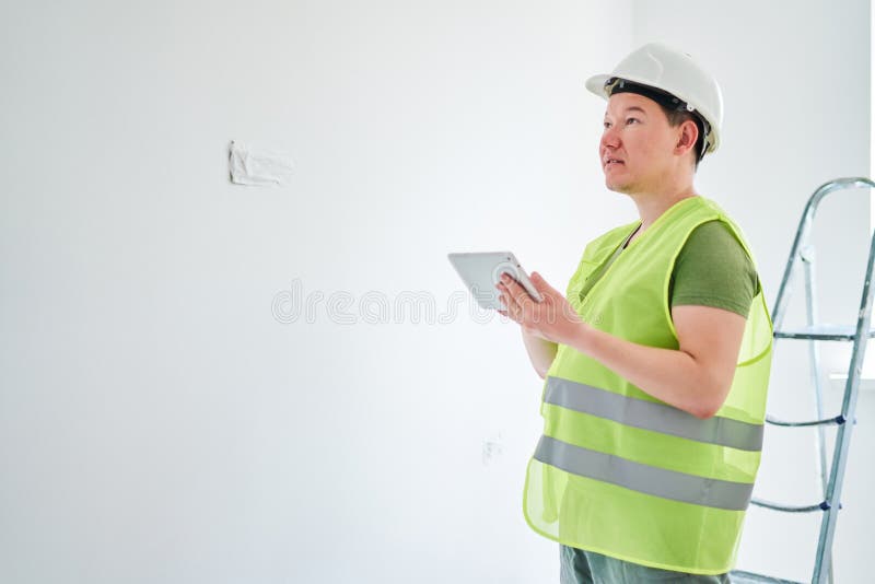 Reparaties in het appartement, een man in een groen vest en een witte helm is in een witte kamer met een tablet in zijn handen stock foto Reparaties in het appartement, een man in een groen vest en een witte helm is in een witte kamer met een tablet in zijn handen stock foto