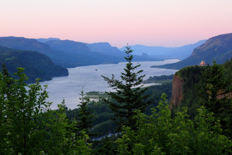 Beautiful evening glow over the Columbia River Gorge. Green trees in forefront, Vista House on the cliff, Beacon Rock in the distance, sweeping majestic Columbia River, territorial mountains of the Cascade Mountain Range, with a glow in the sky in the distance. Beautiful evening glow over the Columbia River Gorge. Green trees in forefront, Vista House on the cliff, Beacon Rock in the distance, sweeping majestic Columbia River, territorial mountains of the Cascade Mountain Range, with a glow in the sky in the distance.