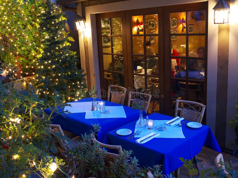 Two tables set in the restaurant patio with blue and white table cloths waiting for diners. Large window showing the the restaurant interior and many Christmas lights on the plants around. Two tables set in the restaurant patio with blue and white table cloths waiting for diners. Large window showing the the restaurant interior and many Christmas lights on the plants around.