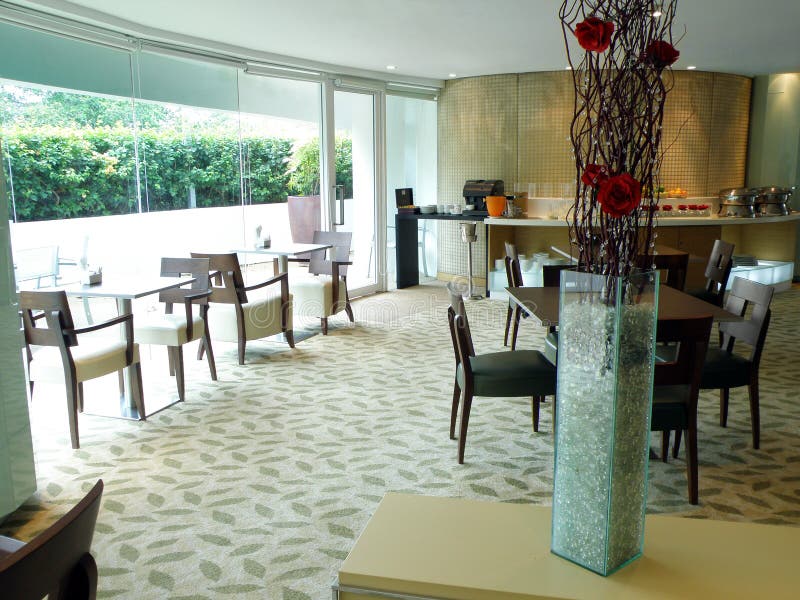 A view of the layout and interior decor of a five star luxurious hotel buffet restaurant dining area. Taken when empty. Located in the contemporary style executive lounge of the resort. Semi natural daylight lighting with large glass walls french windows. With carpets, cool modern floral arrangement, glass and wooden furnitures, etc. Buffet area at far end of room. Simple, subtle and elegant decor theme, colors in red, gold, blue and green. Flower arrangement in large glass vase filled with blue green pebbles. Horizontal color format image. Nobody in picture. Southeast asia location. A view of the layout and interior decor of a five star luxurious hotel buffet restaurant dining area. Taken when empty. Located in the contemporary style executive lounge of the resort. Semi natural daylight lighting with large glass walls french windows. With carpets, cool modern floral arrangement, glass and wooden furnitures, etc. Buffet area at far end of room. Simple, subtle and elegant decor theme, colors in red, gold, blue and green. Flower arrangement in large glass vase filled with blue green pebbles. Horizontal color format image. Nobody in picture. Southeast asia location.