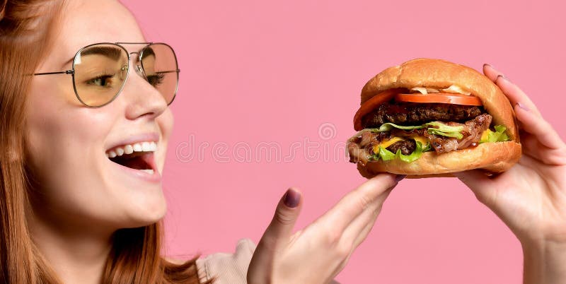 Retrato ascendente cercano de una mujer joven hambrienta que come la hamburguesa sobre fondo rosado fotografía de archivo Retrato ascendente cercano de una mujer joven hambrienta que come la hamburguesa sobre fondo rosado fotografía de archivo