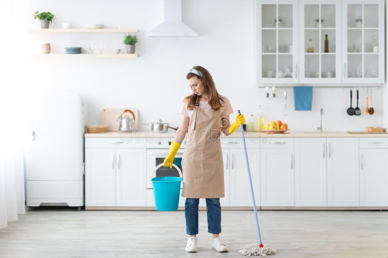 Full length portrait of upset millennial lady with mop and bucket unwilling to wash floor in kitchen, copy space. Millennial housewife sick and tired of cleaning her apartment. Full length portrait of upset millennial lady with mop and bucket unwilling to wash floor in kitchen, copy space. Millennial housewife sick and tired of cleaning her apartment