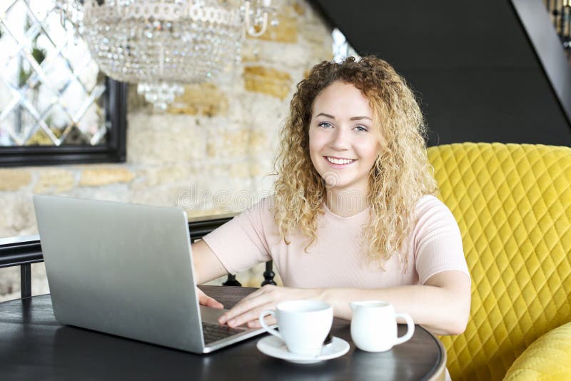 Retrato da jovem mulher loura atrativa, sentando-se na cafetaria, datilografia de sorriso no portátil da tela vazia A fêmea loura fotografia de stock Retrato da jovem mulher loura atrativa, sentando-se na cafetaria, datilografia de sorriso no portátil da tela vazia A fêmea loura fotografia de stock