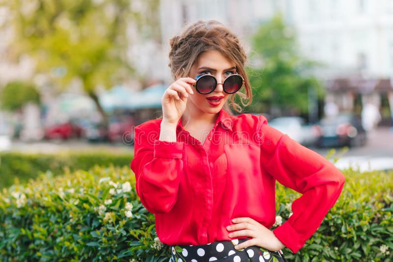 Retrato da menina bonita nos óculos de sol que levantam à câmera no parque Tem o penteado, blusa vermelha fotografia de stock