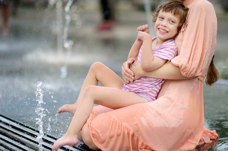 Retrato da menina deficiente bonita nos braços de sua mãe que tem o divertimento na fonte do parque público no dia de verão ensol foto de stock Retrato da menina deficiente bonita nos braços de sua mãe que tem o divertimento na fonte do parque público no dia de verão ensol foto de stock